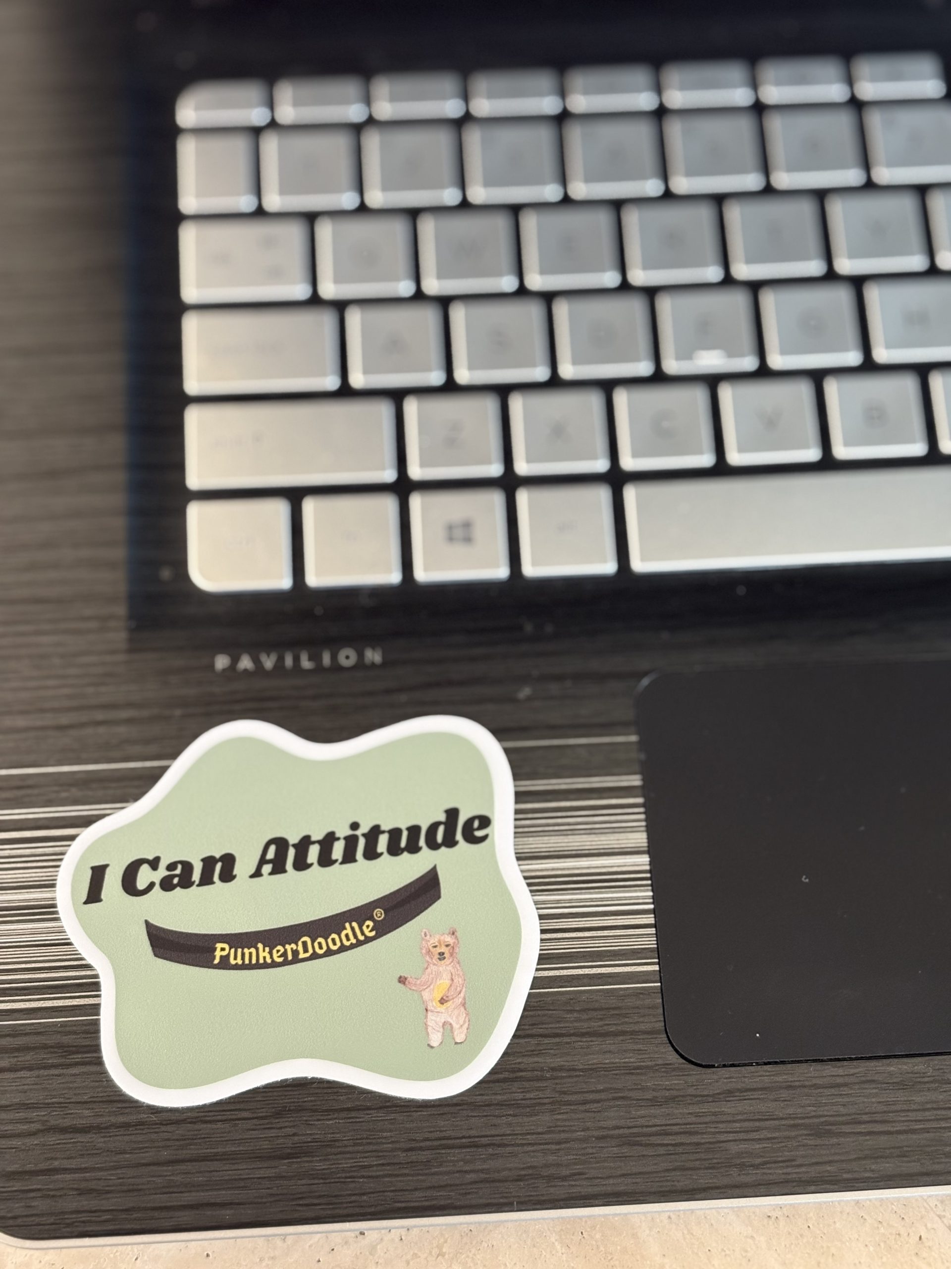 Die-cut I Can Attitude® PunkerDoodle® sticker with JJ Bear on a green background and sticker is placed on laptop.